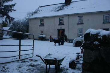 A snowy courtyard in front of a house; a wheelbarrow, stone wall, metal gate, car, and people with a dog create a winter evening scene.
