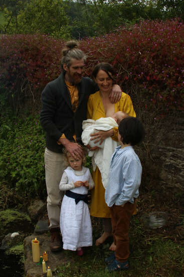 A family of five stands outdoors by a lush, flowering hedge. The mother breastfeeds a baby, while two children and the father lovingly gather close. Candles burn nearby.