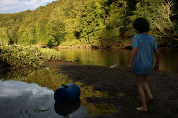 A child in a blue shirt stands barefoot by a riverbank, near a blue barrel, surrounded by lush green trees under a partly cloudy sky.