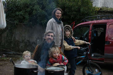 A family of five gathers outdoors beside steaming pots and a red car. Two children sit, one stands with a bicycle. They all wear warm clothes.