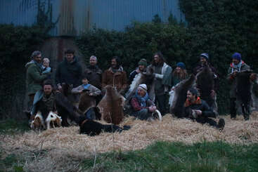 A group of people, including children and dogs, pose outdoors on hay. Many are holding animal hides. They are dressed warmly, standing against a leafy wall.