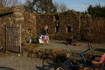 A young girl in a colorful skirt stands beside stone ruins, surrounded by garden tools, plants, a metal gate, and an old blue go-kart under sunlight.