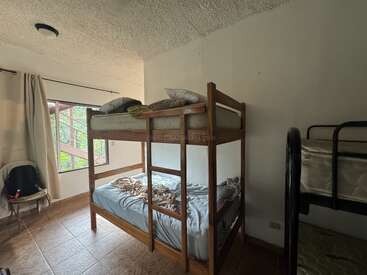 Esta imagen muestra una habitación sencilla con literas de madera, una ventana con cortinas, suelo de baldosas, una silla y una mochila, creando un ambiente similar al de un albergue.