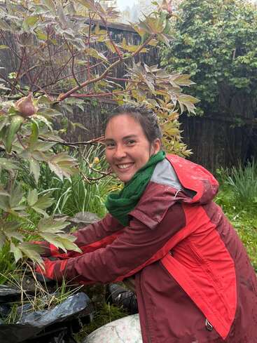Uma mulher sorridente, usando uma capa de chuva vermelha e um cachecol verde, cuida de um jardim, cercada por vegetação e plantas. Parece ser um dia chuvoso.