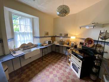 La imagen muestra una cocina con ventana, fregadero, fogones y encimeras, con suelo a cuadros y varios electrodomésticos, en un espacio bien iluminado y organizado.