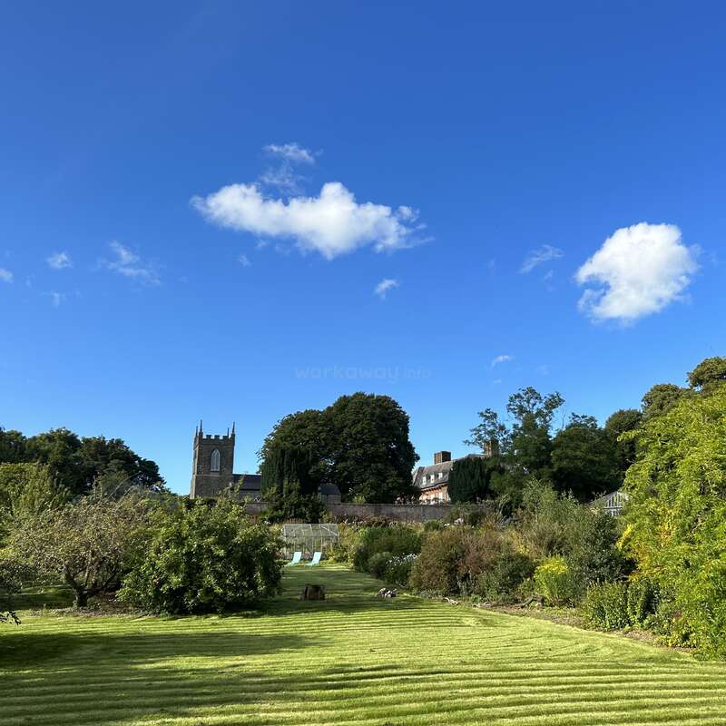 La imagen muestra un paisaje sereno con un césped verde, árboles, arbustos y la torre de una iglesia a lo lejos, sobre un cielo azul despejado con algunas nubes.
