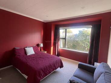 Esse quarto tem paredes vermelhas arrojadas, uma cama aconchegante com roupa de cama combinando, uma janela com cortinas, uma mesa lateral, luminária e um confortável sofá cinza.