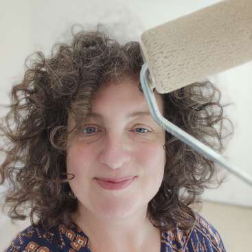 A woman with curly brown hair smiles at the camera, holding a paint roller. She appears to be painting, standing in a brightly lit, neutral room.