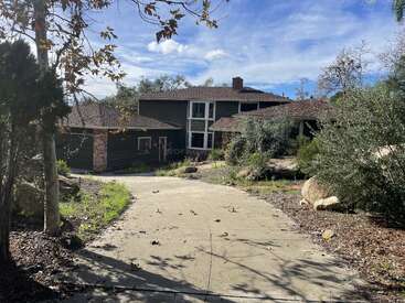 A two-story house with a brick chimney sits at the end of a sloped driveway, surrounded by trees, rocks, and natural landscaping under a bright sky.