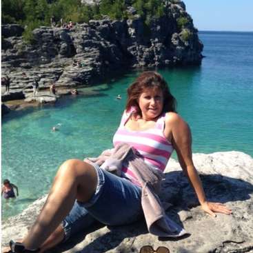 La imagen muestra a una mujer sentada en un peñasco, con una masa de agua y un acantilado al fondo, rodeada de gente nadando y tomando el sol.