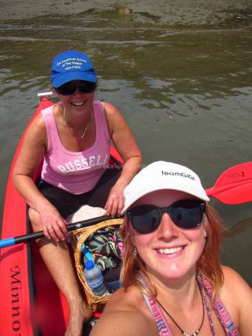 La imagen muestra a dos mujeres en una canoa sobre el agua, una de ellas con un remo en la mano y la otra con un sombrero blanco.
