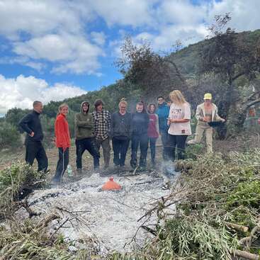 L'image représente un groupe de personnes autour d'un feu dans une zone boisée, entourée d'arbres et d'un ciel bleu avec des nuages blancs.