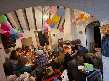 A group of people gather around a festive table in a cozy room decorated with colorful balloons, enjoying food, drinks, and conversation around a warm fireplace.