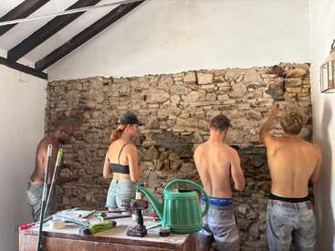 Four people are working together to restore or renovate a rustic stone wall indoors. Tools, cleaning supplies, and a green watering can are on a nearby table.