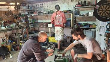 Three men are working in a cluttered garage filled with tools, shelves, and equipment. One is standing, two are fixing something, and a dog is resting nearby.
