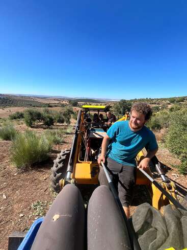 A man in a blue shirt rides a yellow tractor in a rural, hilly landscape with bright blue skies. Others are seated behind him, enjoying the view.