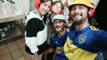 Four smiling people pose together indoors, wearing red ear protection. Two men wear blue Boca Juniors jerseys. Shelves with bottles and jugs are visible in the background.