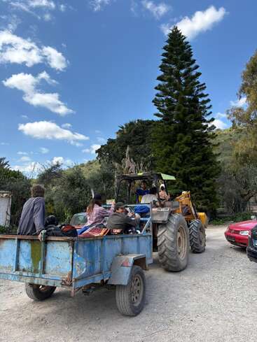 Um grupo de pessoas viaja em um reboque azul acoplado a um trator, cercado por árvores, carros e um céu limpo com algumas nuvens espalhadas.