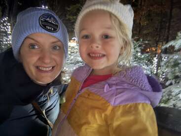 Une femme et une jeune fille sourient chaleureusement à l'appareil photo, vêtues de vestes et de bonnets d'hiver, entourées de lumières de Noël scintillantes et d'arbres givrés dans la nuit.