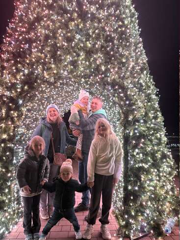 Une famille heureuse pose sous une grande arche de sapin de Noël magnifiquement éclairée la nuit, entourée de lumières scintillantes, de sourires et de vêtements d'hiver festifs, créant ainsi de joyeux souvenirs.