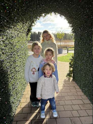 Quatre enfants se tiennent sous une arche festive décorée de lumières, en plein air par une journée ensoleillée, souriant et posant pour une joyeuse photo d'hiver ou de vacances.