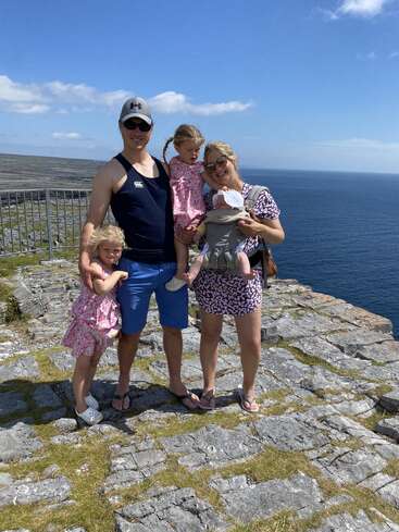 L'image représente une famille de cinq personnes posant sur une falaise rocheuse surplombant l'océan, avec un ciel bleu serein et des eaux calmes en arrière-plan.