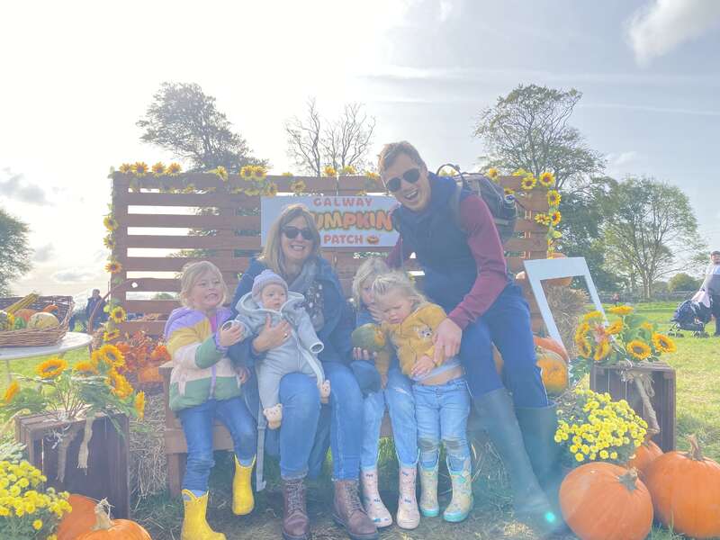 L'image représente une famille de cinq personnes posant pour une photo dans un champ de citrouilles, entourée de citrouilles et de tournesols, avec un panneau indiquant "Galway Pumpkin Patch" à l'arrière-plan.