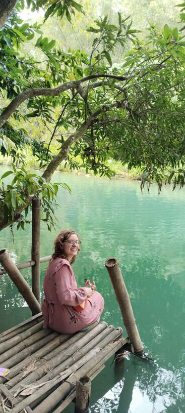 A woman in a pink dress sits on a bamboo dock by a clear, greenish lake, smiling, surrounded by lush trees and peaceful nature.
