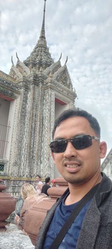 A man wearing sunglasses takes a selfie in front of an ornate, traditional Asian temple. Other tourists are visible, and the sky is filled with clouds.