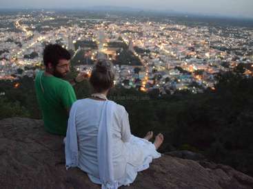 Un hombre y una mujer sentados en un peñasco con vistas a una ciudad al atardecer, con las luces de la ciudad parpadeando bajo ellos.