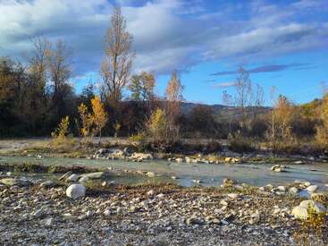 A peaceful autumn landscape with a shallow river, rocks, and trees in golden-yellow foliage under a partly cloudy blue sky, capturing a tranquil natural scene.