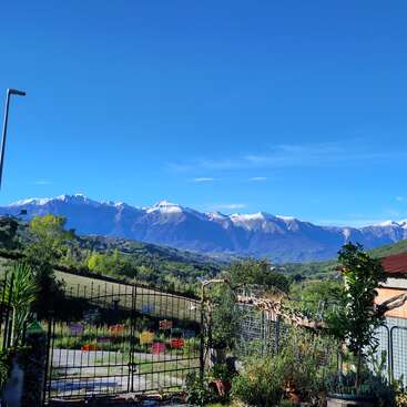 A stunning landscape features a garden gate opening to lush green hills, vibrant plants, and majestic snow-capped mountains under a clear, bright blue sky.
