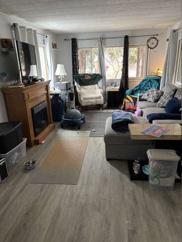 A cozy living room with a fireplace, TV, sectional sofa, chairs, yoga mat, and scattered pet toys. Large windows bring in natural light. Lived-in feel.