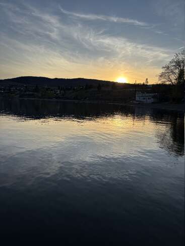L'image montre un lac paisible au coucher du soleil, avec le soleil qui se couche derrière des collines lointaines, se reflétant sur l'eau calme, et une maison dont la silhouette se détache parmi les arbres.