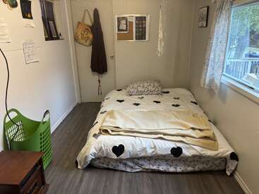 Une chambre intime et épurée, avec un matelas posé à même le sol, une parure de lit à motifs de cœurs, une fenêtre ornée de voilages, un panier à linge, des sacs suspendus à des crochets et un tableau d'affichage.