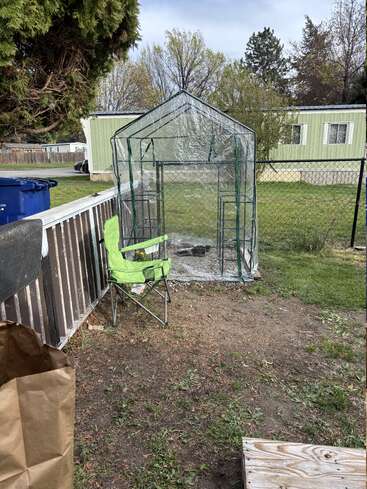 Une petite serre recouverte de plastique transparent se trouve dans un jardin. À proximité se trouvent une chaise pliante verte, un sac en papier pour les déchets verts et un escabeau en bois.