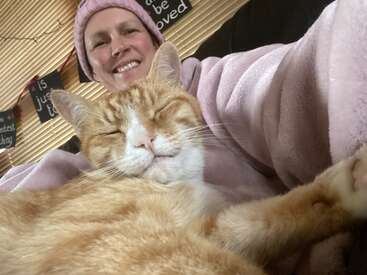 The image shows a woman in a pink beanie and robe sitting with an orange and white cat on her lap, both looking at the camera.