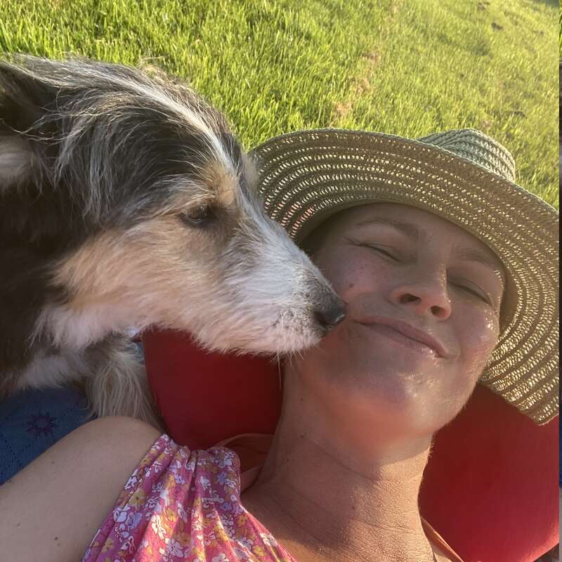The image depicts a serene scene of a woman reclining on a red cushion in a lush grassy field, with a dog tenderly nuzzling her face.