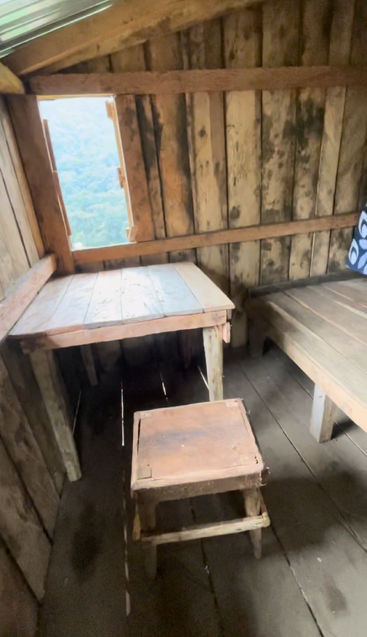 Esta imagen muestra una habitación rústica de madera con una pequeña mesa, un taburete sencillo, un banco, paredes de madera y una ventana que revela una vista verde del bosque.