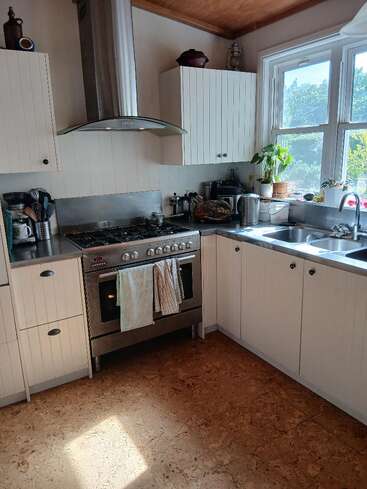 Bright, cozy kitchen featuring white cabinets, stainless steel oven, range hood, double sink, potted plant, large windows, wooden ceiling, cork flooring, and countertop appliances.