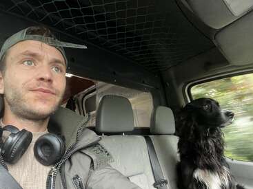 Un homme avec des écouteurs autour du cou et une casquette à l'envers est assis dans un véhicule à côté d'un chien noir et blanc qui regarde par la fenêtre.