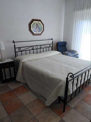 En esta imagen se ve una cama bien hecha con estructura metálica, una colcha acolchada, una mesita de noche con una lámpara, un sillón, suelo de baldosas y una obra de arte religiosa.