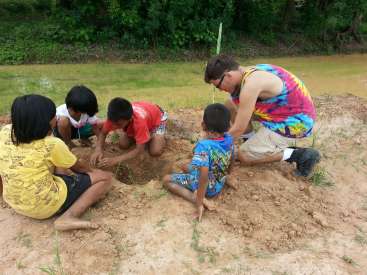 The image depicts a man and four children digging a hole in the dirt, with the man wearing a tie-dye tank top and the children in casual clothing.