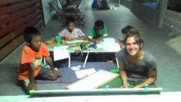 The image depicts a woman and three children seated on the floor, engaged in a creative activity with art supplies and papers.
