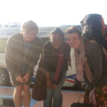La imagen muestra a tres individuos posando delante de un autobús: el hombre de la izquierda lleva pantalones cortos y chaqueta, la mujer del centro lleva bufanda y gafas, y la mujer de la derecha lleva una mochila.