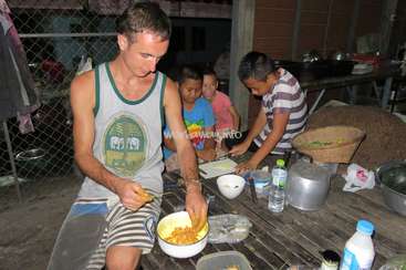 The image depicts a man and three children preparing food in a rustic outdoor setting, with a wooden table and various cooking utensils and ingredients visible.