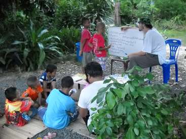 The image depicts a man teaching a group of children in an outdoor setting, utilizing a whiteboard and a blue plastic chair. The children are seated on the ground.