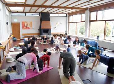 Un grupo de adultos y niños practican yoga juntos en una sala luminosa y espaciosa con grandes ventanales, techo de madera y chimenea. Las esterillas rodean un altar central.