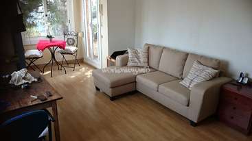La imagen muestra un salón con un sofá beige, una mesa de madera y un balcón con un mantel rojo. La habitación está bien iluminada y tiene una combinación de colores neutros.