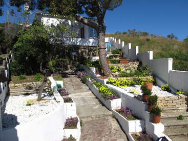 La imagen muestra una serena casa blanca con un camino de piedra y escaleras que conducen a ella, rodeada de un hermoso jardín con flores vibrantes y exuberante vegetación.